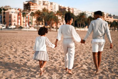 Beyaz elbiseli, tatlı kıvırcık bir kızı olan Afro-Amerikan bir aile şafak vakti kumlu bir sahilde sahil evlerinin önünde yürüyor ve eğleniyorlar..