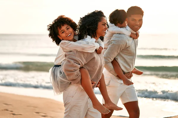 African American family with a cute curly daughter in a white dress and a little son on their backs walking and having fun on the sandy beach by the sea at dawn.