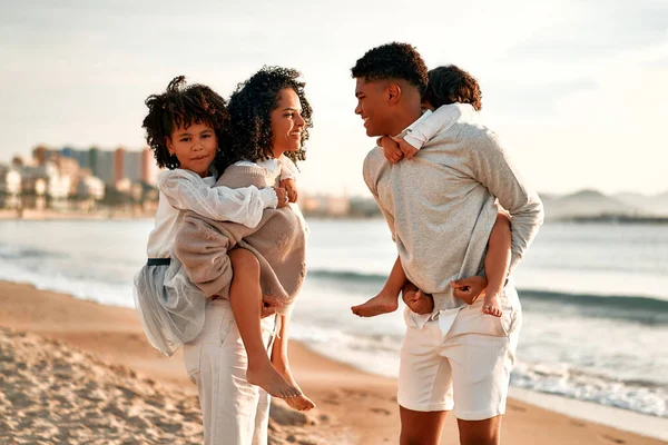 白いドレスを着たかわいい娘と背中の小さな息子を持つアフリカ系アメリカ人の家族が歩くと夜明けに海で砂浜で楽しみます — ストック写真