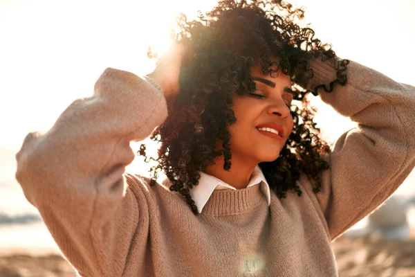 Beautiful African American woman with afro curls in a warm sweater sitting on a sandy beach near the sea, looking at the seascape.