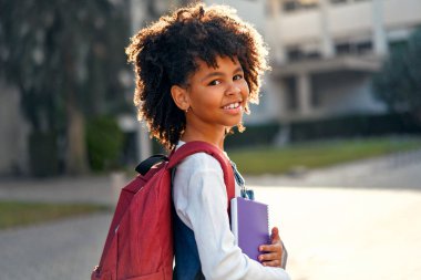 Kıvırcık saçlı, okul çantalı ve elinde not defterleri olan mutlu Afro-Amerikan akıllı kızı. Modern sırt çantası. Okula ilk gelişim. Okula geri dön..