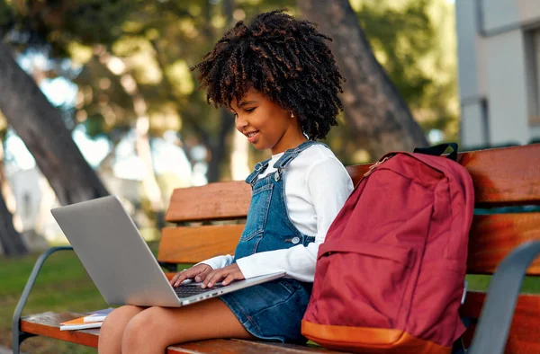 Back to school. A cute African American child girl with afro hairstyle with a backpack sitting on a bench near the school with a laptop. Primary school student after classes learning homework.