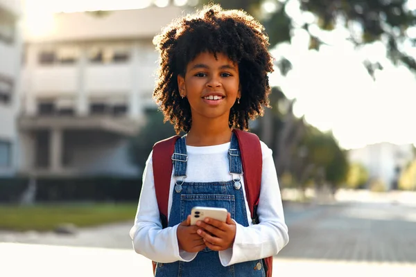 Back to school. Happy cute African American smart girl with backpack holding mobile phone playing with mobile phone outdoors. Schoolgirl pupil with a bag. Elementary school student after class.