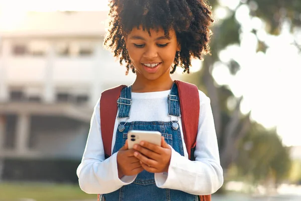Back to school. Happy cute African American smart girl with backpack holding mobile phone playing with mobile phone outdoors. Schoolgirl pupil with a bag. Elementary school student after class.