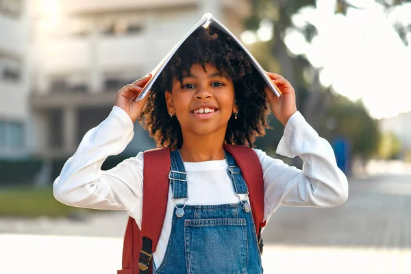 Back to school. A cute African American schoolgirl with a backpack made a roof on her head from a book, standing on the street against the backdrop of the school.