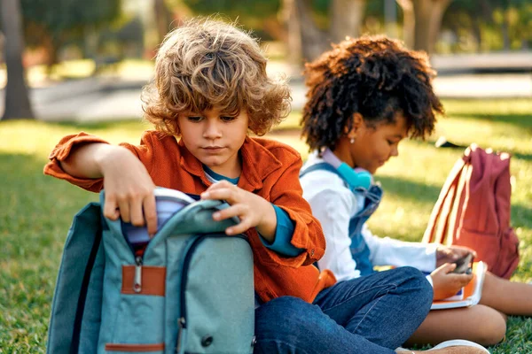 Charming schoolchildren after lessons are resting sitting on the grass in the park. Back to school. African American girl with a Caucasian boy take out a lunch box from their backpack to have lunch.