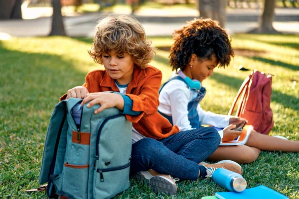 Charming schoolchildren after lessons are resting sitting on the grass in the park. Back to school. African American girl with a Caucasian boy take out a lunch box from their backpack to have lunch.