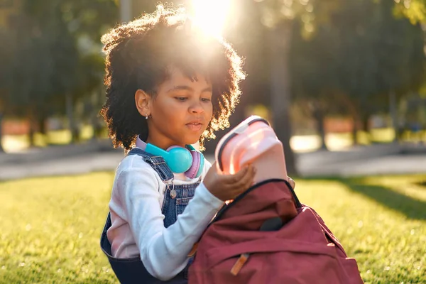 迷人的非洲裔美国女学生课后坐在公园的草地上休息 回学校去一个非洲裔美国女孩从背包里拿出一个午餐盒来吃午饭 — 图库照片