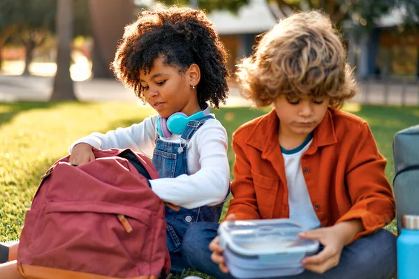 课后迷人的学童正坐在公园的草地上休息 回学校去和白人男孩在一起的非洲裔美国女孩从背包里拿出一个午餐盒来吃午饭 — 图库照片