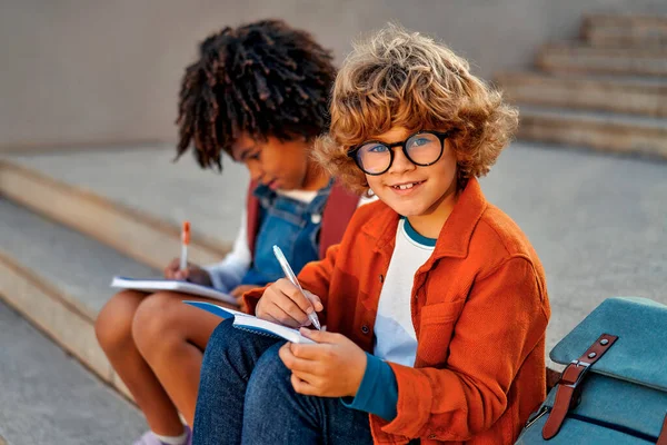 聪明的学童坐在学校附近的楼梯上一起做作业很迷人 回学校去与戴眼镜的白人男孩一起学习的非裔美国女孩 — 图库照片