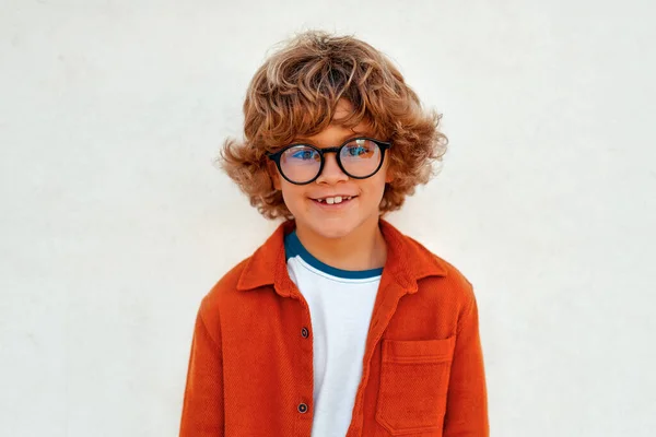 Charming smart child with a stylish hairstyle in glasses smiling while standing against the background of a white wall. School concept.