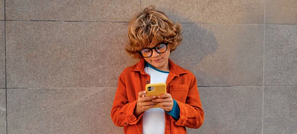 Charming smart child with a stylish hairstyle in glasses with a phone standing against the background of a wall. School concept.