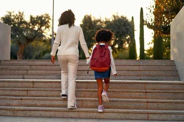 Happy African American mother and daughter preschool student walking to school. Beginning of lessons. First day of fall. Parenthood or love and bonding expression concept.