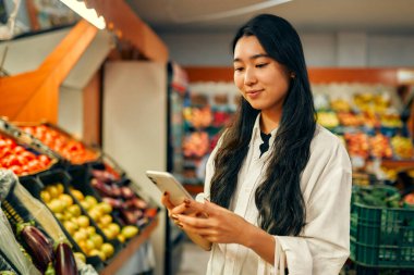 Sebze ve meyve dükkanında akıllı telefon kullanan Asyalı genç bir kadın. Sağlıklı yiyecekler. Eve yemek teslimatı. İnternetten alışveriş.