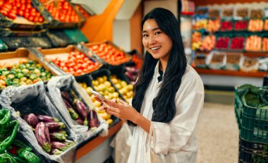 Sebze ve meyve dükkanında akıllı telefon kullanan Asyalı genç bir kadın. Sağlıklı yiyecekler. Eve yemek teslimatı. İnternetten alışveriş.