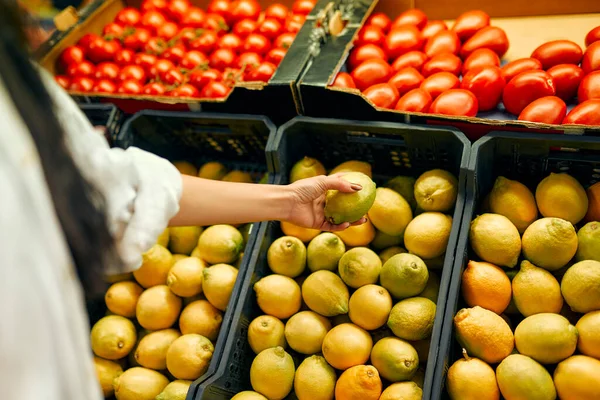 A young woman chooses lemons in a store. Buying fresh vegetables and fruits. Healthy food. Crates of lemons and tomatoes are for sale.