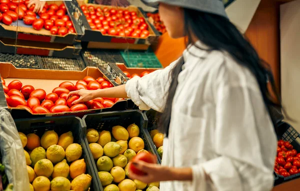 A young Asian woman chooses tomatoes in a store. Buying fresh vegetables and fruits. Healthy food. Crates of lemons and tomatoes are for sale.