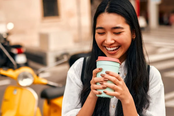 Portrait of an Asian female tourist student in a white shirt with a backpack drinks morning coffee while standing against the background of a scooter on the city streets. Vacation and tourism concept.