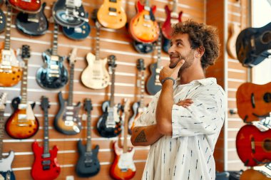 Handsome bearded curly man standing in front of walls with many electric guitars in a musical instrument store and choosing which one to buy. Hobbies and recreation. Buying a guitar in a store.