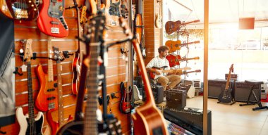 Arka planda sakallı yakışıklı kıvırcık adam bir müzik aletleri mağazasında bir sürü elektrikli gitarla duvara yaslanmış ve çalmaya çalışıyor. Hobiler ve eğlence. Bir mağazadan gitar almak..