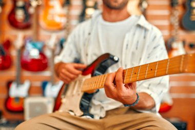 Handsome bearded curly man in front of walls with many electric guitars in a musical instrument store and trying to play it. Hobbies and recreation. Buying a guitar in a store.