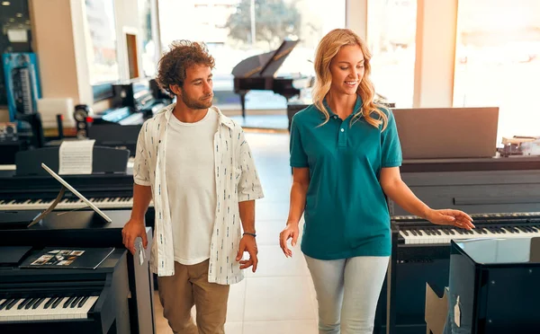 Handsome bearded curly-haired man choosing a piano in a musical instrument store getting the help of a consultant. Woman seller telling the buyer about the piano.
