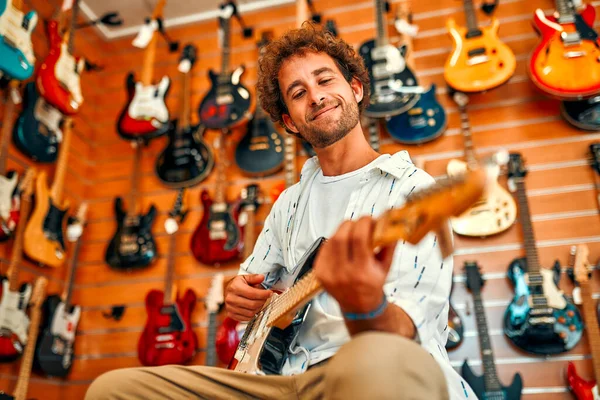 Handsome bearded curly man in front of walls with many electric guitars in a musical instrument store and trying to play it. Hobbies and recreation. Buying a guitar in a store.