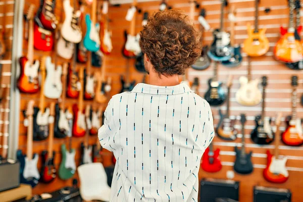 Handsome bearded curly man standing in front of walls with many electric guitars in a musical instrument store and choosing which one to buy. Hobbies and recreation. Buying a guitar in a store.