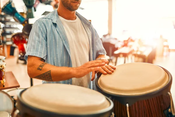 Handsome bearded curly man playing drums with his hands in a musical instrument store before buying.