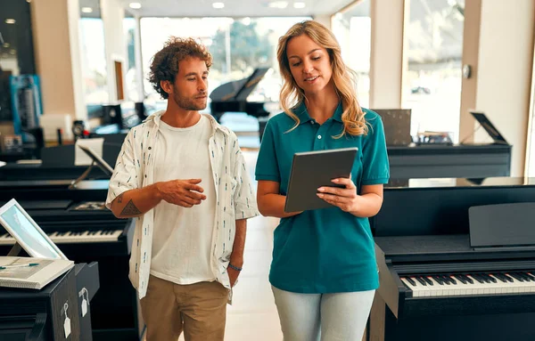 Handsome bearded curly-haired man choosing a piano in a musical instrument store getting the help of a consultant. Woman seller telling the buyer about the piano.