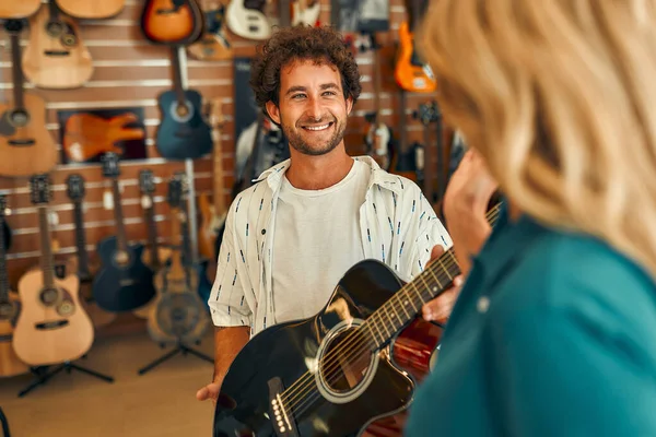 Handsome bearded curly man choosing a guitar in a musical instrument store getting the help of a consultant. Woman seller telling about assortment of guitars to buyer.