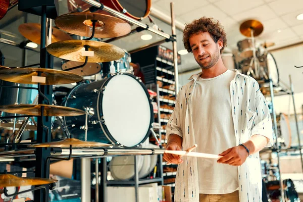 Handsome bearded curly man choosing a drum in a musical instrument store. Hobbies and recreation. A man buying a drum in a store and trying to play it.