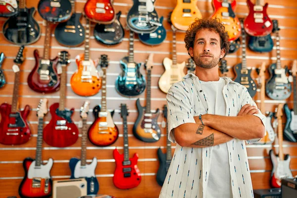 Handsome bearded curly man standing in front of walls with many electric guitars in a musical instrument store and choosing which one to buy. Hobbies and recreation. Buying a guitar in a store.