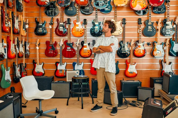 Handsome bearded curly man standing in front of walls full of electric guitars in a musical instrument store. Hobbies and recreation. Buying a guitar in a store.