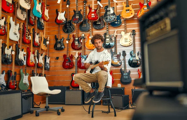 Handsome bearded curly man in front of walls with many electric guitars in a musical instrument store and trying to play it. Hobbies and recreation. Buying a guitar in a store.