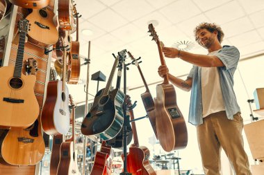 Handsome bearded curly man choosing a guitar in a musical instrument store. Hobbies and recreation.