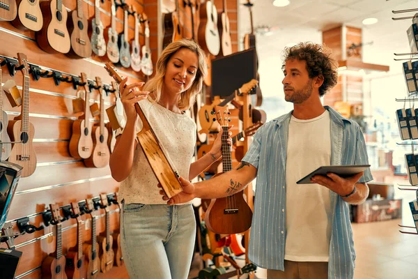 A young blonde woman buying a ukulele guitar asking for help from a sales assistant in a musical instrument store against the backdrop of walls with many different guitars.