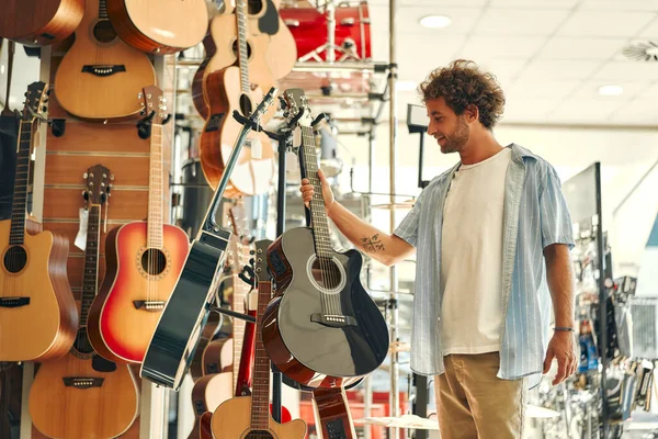 Handsome bearded curly man choosing a guitar in a musical instrument store. Hobbies and recreation.