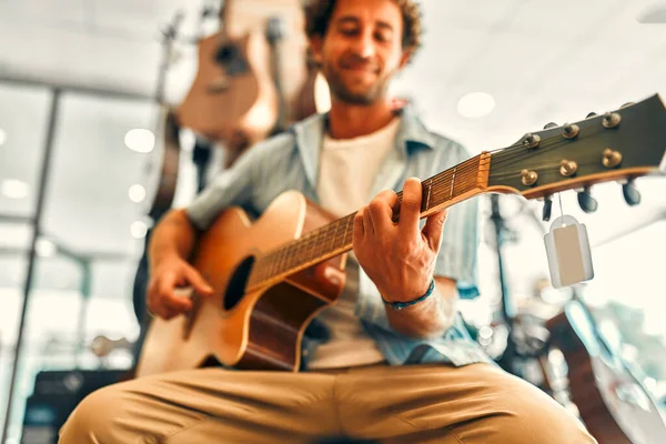 A handsome bearded curly man chooses a guitar in a musical instrument store and trying to play it. Hobbies and recreation. Buying a guitar in a store.