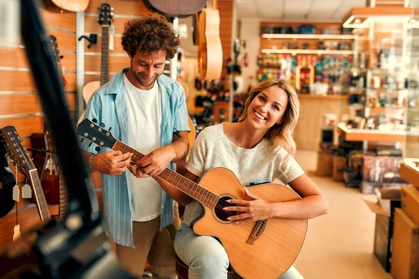 Beautiful young woman choosing a guitar in a musical instrument store and trying to play it with the help of a sales assistant male. Young couple buying a guitar in a store.