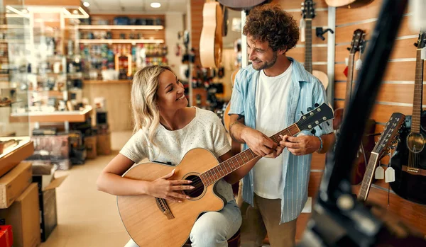 Beautiful young woman choosing a guitar in a musical instrument store and trying to play it with the help of a sales assistant male. Young couple buying a guitar in a store.