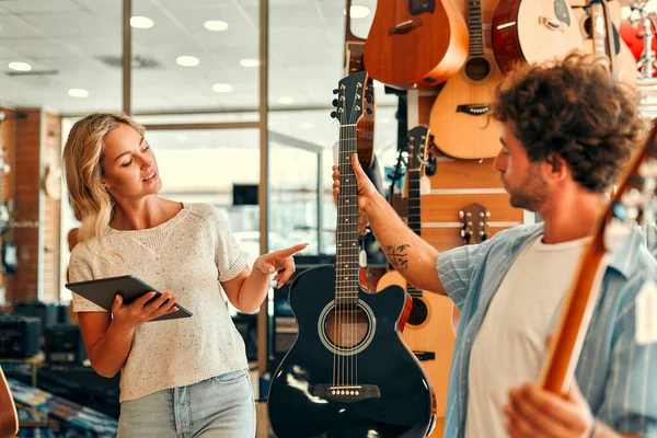 Young bearded male choosing a guitar in a musical instrument store and getting the help of a sales assistant woman.