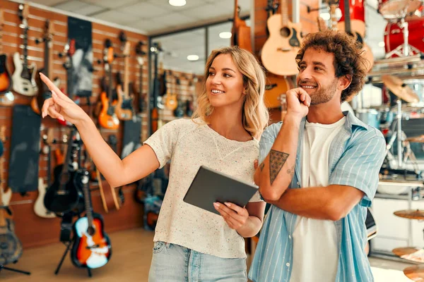 Young female sales assistant helping a customer to choose a guitar in a musical instrument store against the backdrop of walls with many different guitars.
