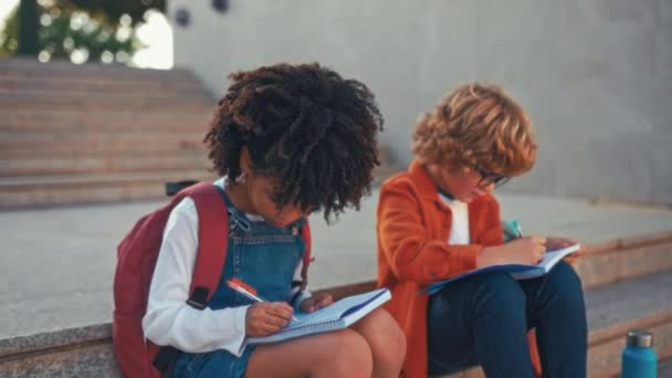 聪明的学童坐在学校附近的楼梯上一起做作业很迷人 回学校去与戴眼镜的白人男孩一起学习的非裔美国女孩 — 图库视频影像