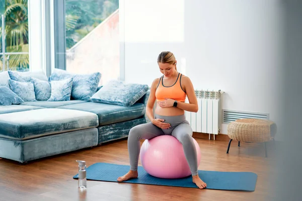 一位穿着运动服的年轻孕妇坐在健身球上和客厅里的橡胶垫子上做运动时 穿着运动服 怀孕期间的运动 — 图库照片