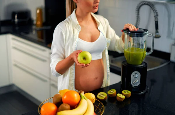 一个年轻的孕妇正在厨房里用搅拌机准备一个冰沙 一个孕妇喝维生素冰沙 怀孕期间健康饮食的概念 — 图库照片