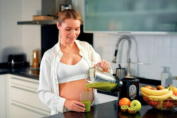 一个年轻的孕妇正在厨房里用搅拌机准备一个冰沙 一个孕妇喝维生素冰沙 怀孕期间健康饮食的概念 — 图库照片