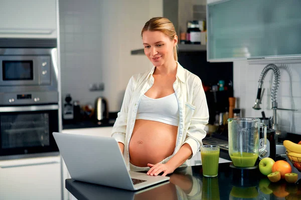 一位年轻的孕妇在厨房里带着笔记本电脑 喝着维生素冰沙 怀孕期间健康饮食的概念 — 图库照片