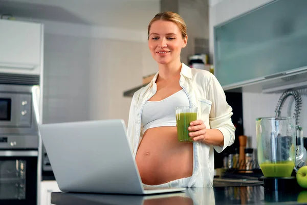 一位年轻的孕妇在厨房里带着笔记本电脑 喝着维生素冰沙 怀孕期间健康饮食的概念 — 图库照片