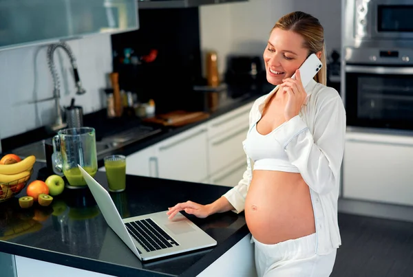 一个年轻的孕妇在厨房里的笔记本电脑前工作 在电话里聊天 在一张刚刚准备好的维生素冰沙的桌子上 怀孕期间的工作和健康饮食 — 图库照片